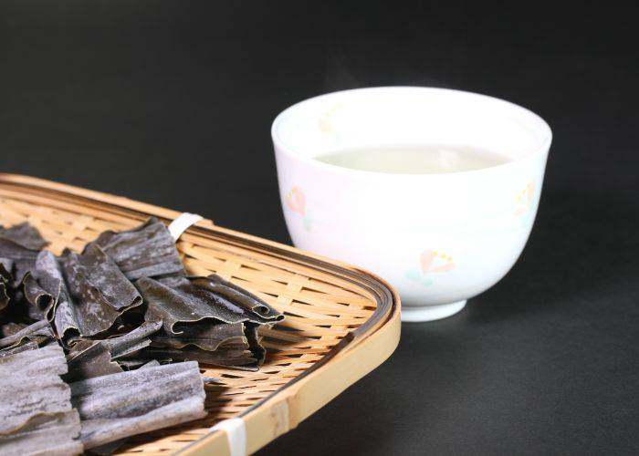 Dried kombu kelp on a bamboo tray, next to a white cup filled with clear kombucha.
